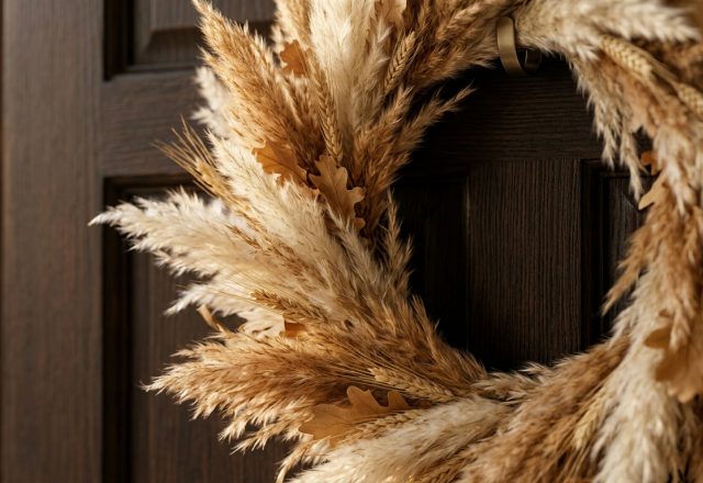 A minimalist fall front door wreath made of pampas grass and wheat hanging on a dark wood door.