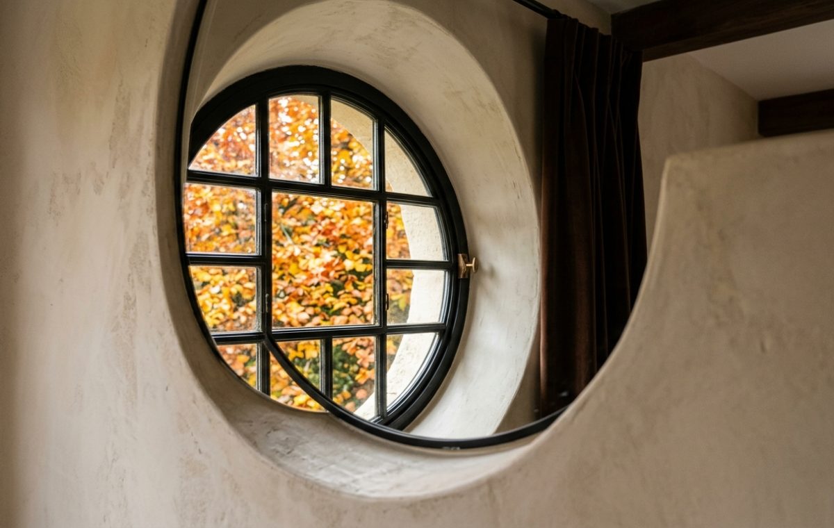 A striking black-framed round window set into a thick plaster wall revealing autumn foliage outside.