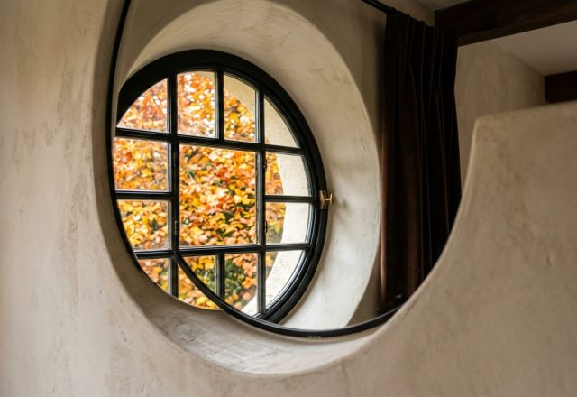 A striking black-framed round window set into a thick plaster wall revealing autumn foliage outside.