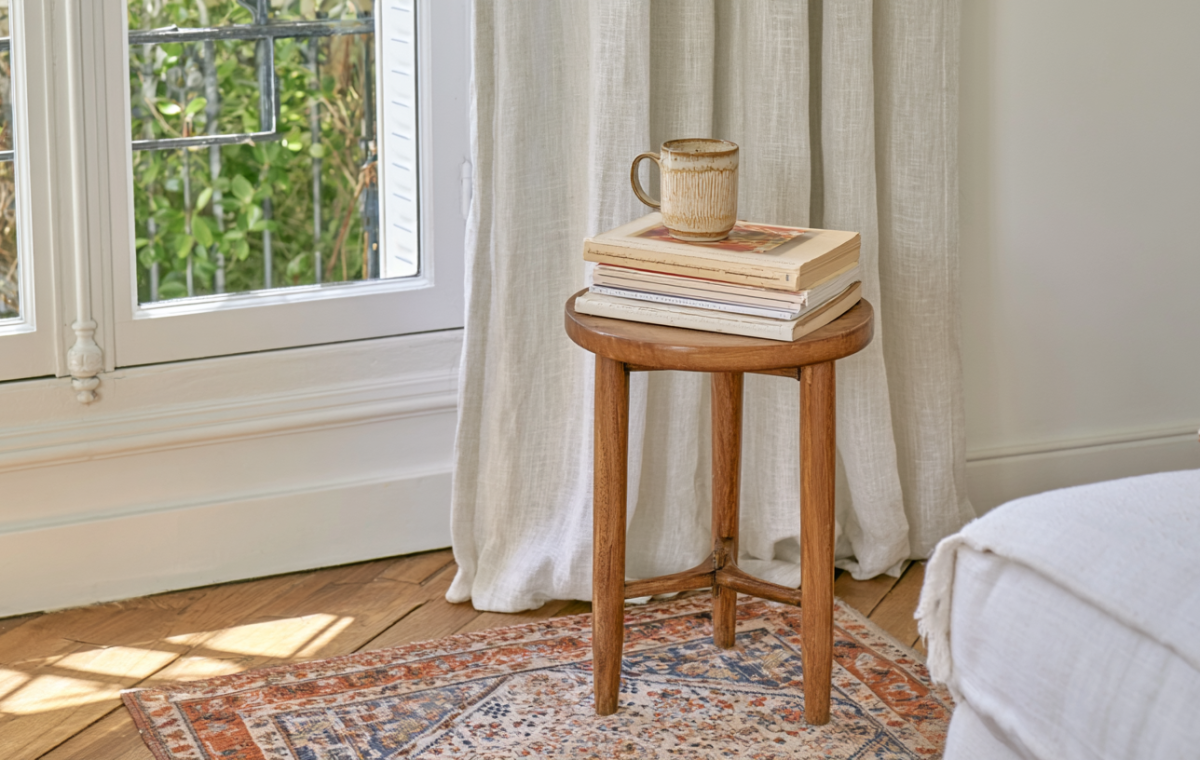 A small wooden side table holding books and a mug next to a window in a cozy living room