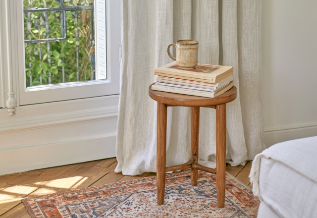 A small wooden side table holding books and a mug next to a window in a cozy living room