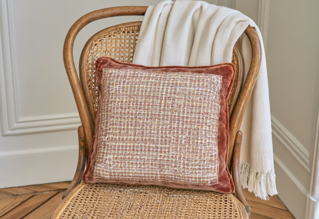 A patterned textured accent pillow resting on a vintage cane back chair with a cream knit blanket.