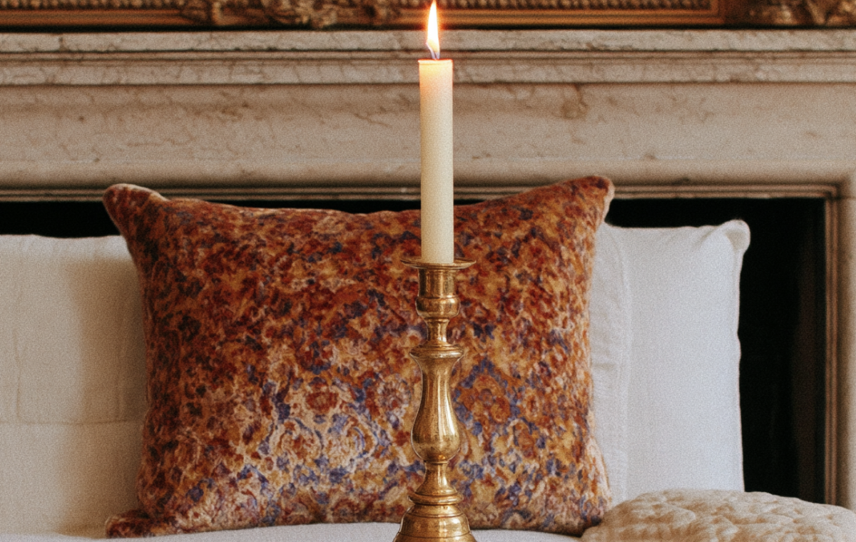 A glowing brass candlestick holder on a mantelpiece with a patterned velvet pillow in a traditional bedroom.