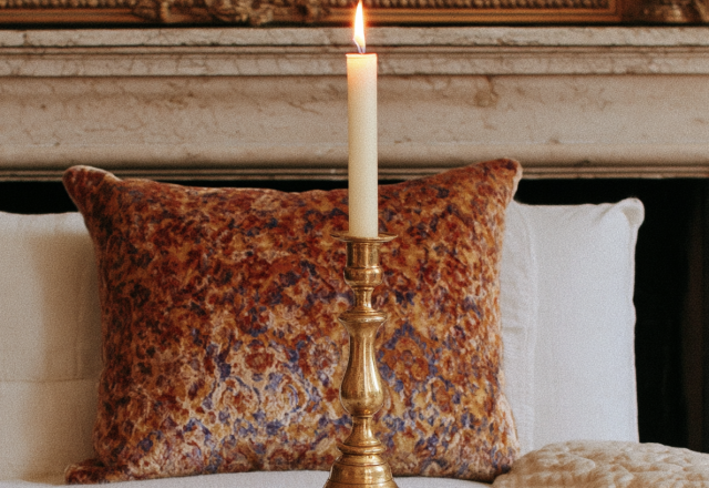 A glowing brass candlestick holder on a mantelpiece with a patterned velvet pillow in a traditional bedroom.