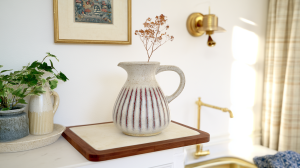 A speckled ceramic jug vase with red stripes styled on a wooden board with dried botanicals in a bright kitchen.