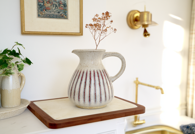 A speckled ceramic jug vase with red stripes styled on a wooden board with dried botanicals in a bright kitchen.