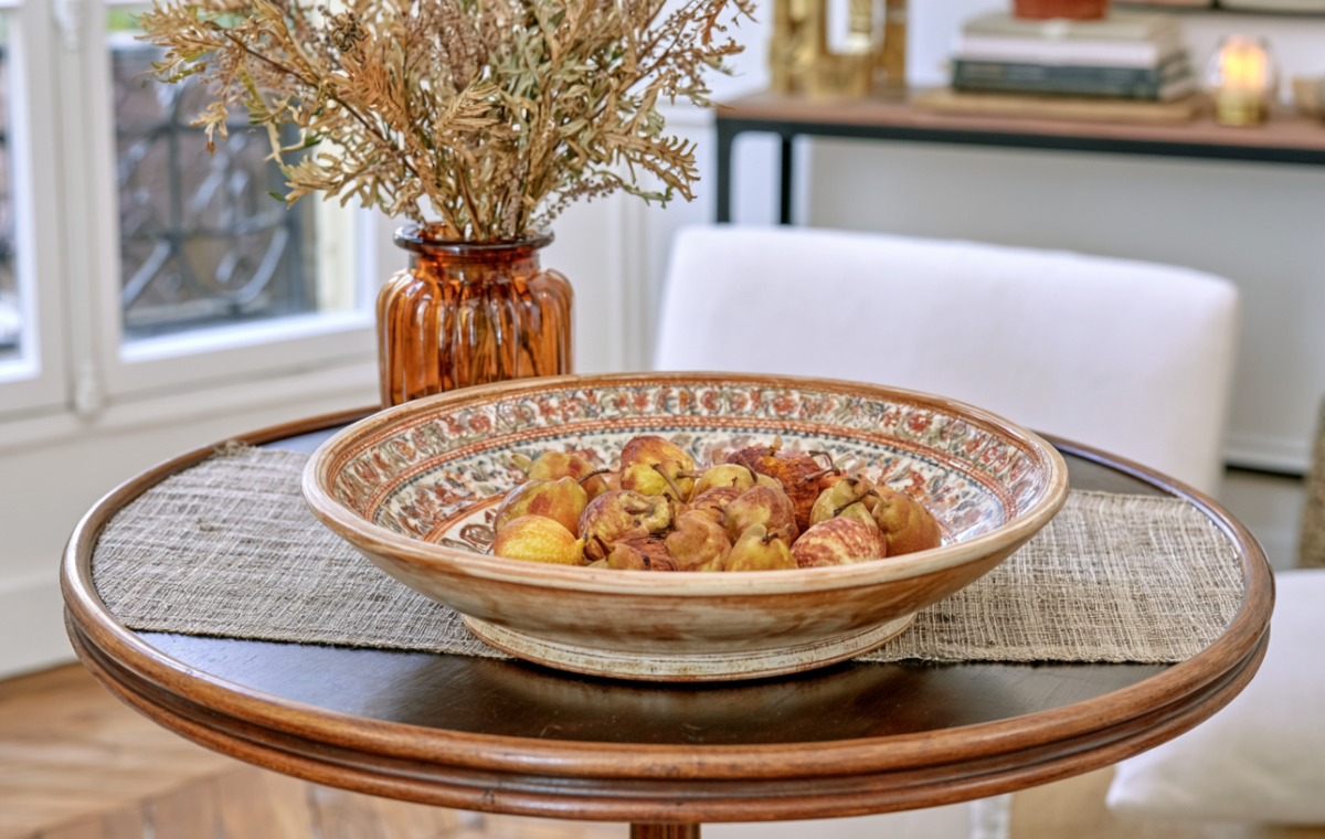 A decorative fruit bowl filled with pears sits on a round wooden table with a textured runner.