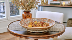A decorative fruit bowl filled with pears sits on a round wooden table with a textured runner.