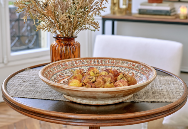 A decorative fruit bowl filled with pears sits on a round wooden table with a textured runner.