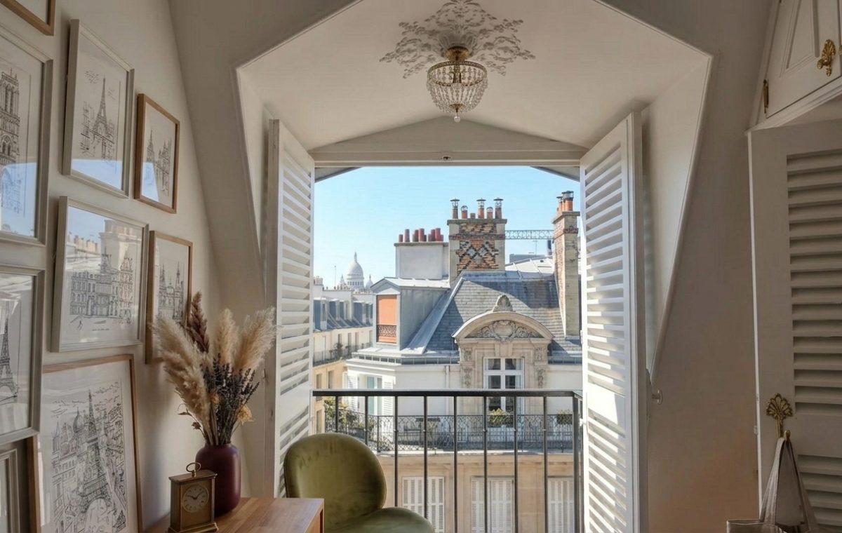 A bright French apartment balcony viewed through an open window with Parisian architecture outside and a cozy study nook inside.