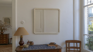 Square French Country mirror mounted on a white wall above a vintage wooden console table with a lamp.