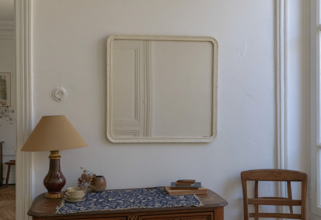 Square French Country mirror mounted on a white wall above a vintage wooden console table with a lamp.