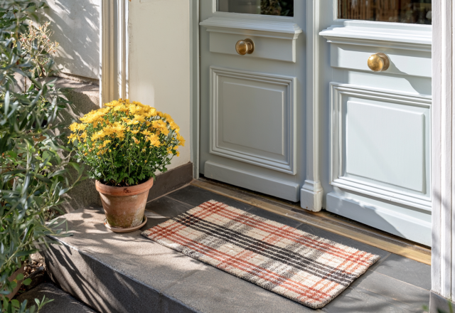 Plaid front entryway doormat on dark stone steps next to potted yellow flowers and light grey double doors.