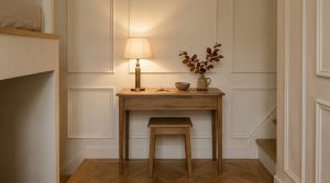 A shallow wooden hallway console desk with matching tucked stool against paneled cream walls.