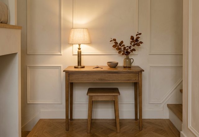 A shallow wooden hallway console desk with matching tucked stool against paneled cream walls.