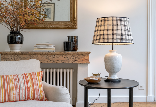 A cozy living room corner featuring a plaid lampshade on a white ceramic table lamp beside a striped throw pillow and marble mantel.