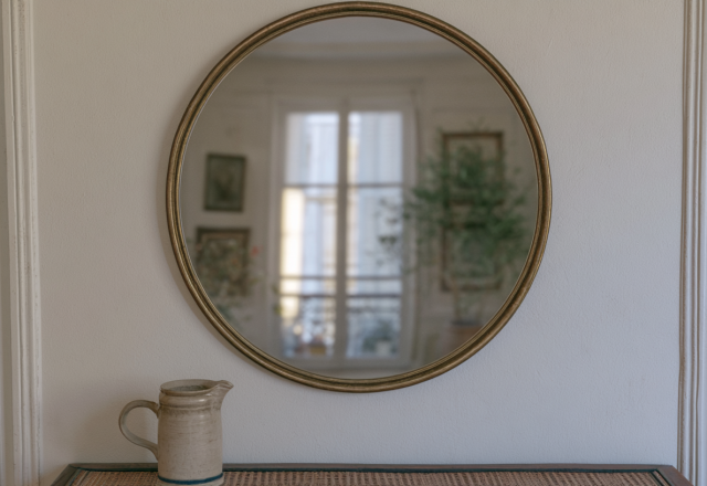 A minimalist round brass mirror hanging above a stoneware pitcher on a textured entryway table.