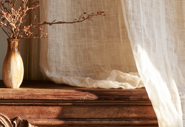 A rustic wooden mantelpiece styled with a ceramic vase of dried branches and a translucent linen curtain.