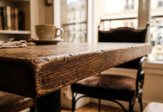 Close-up of a rustic wooden table with a coffee cup and vintage chairs in a sunlit room.
