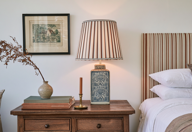 A cozy striped headboard bedroom featuring a rustic wooden nightstand, blue ceramic lamp with a striped shade, and vintage framed art.