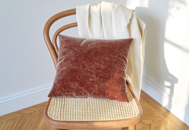 A terracotta velvet accent pillow and cream throw blanket resting on a wooden cane chair in a sunlit room.