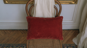 A plush red velvet accent pillow resting on a vintage cane chair against a neutral wall.