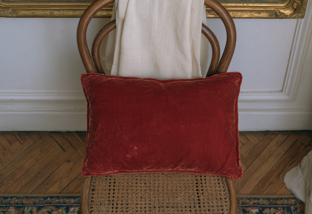 A plush red velvet accent pillow resting on a vintage cane chair against a neutral wall.