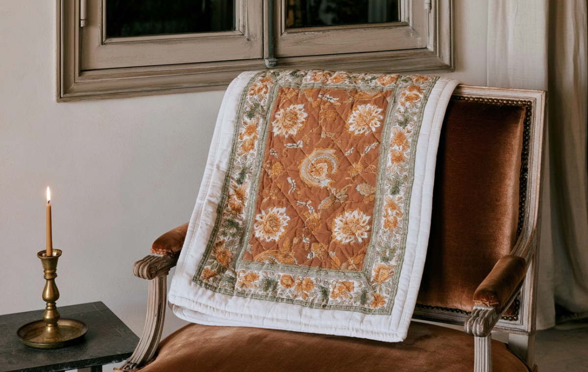 A vintage floral quilt draped over a brown velvet armchair next to a brass candlestick.
