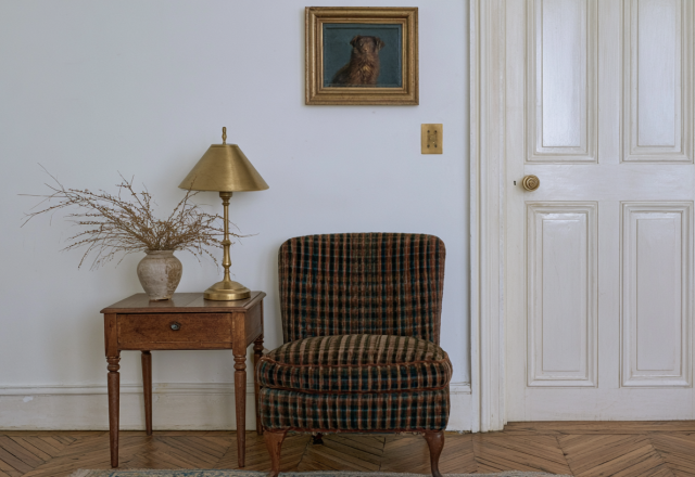 Vintage plaid armchair next to a wooden side table with a brass lamp and framed dog painting.