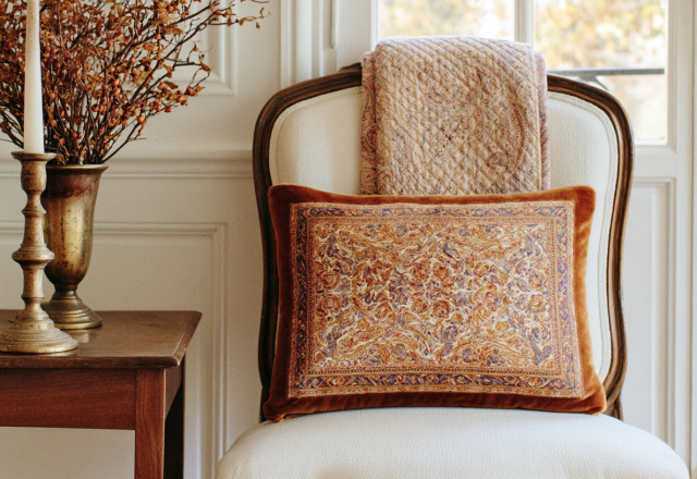 A vintage velvet pillow with intricate patterned fabric resting on a white upholstered antique chair next to a brass candleholder on a wood side table.