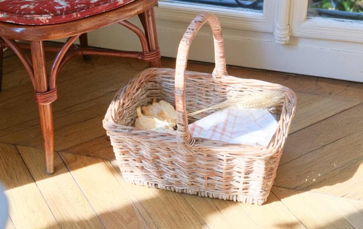 A wicker market basket with linen and dried wheat sitting on sunlit wooden floors next to an antique chair.