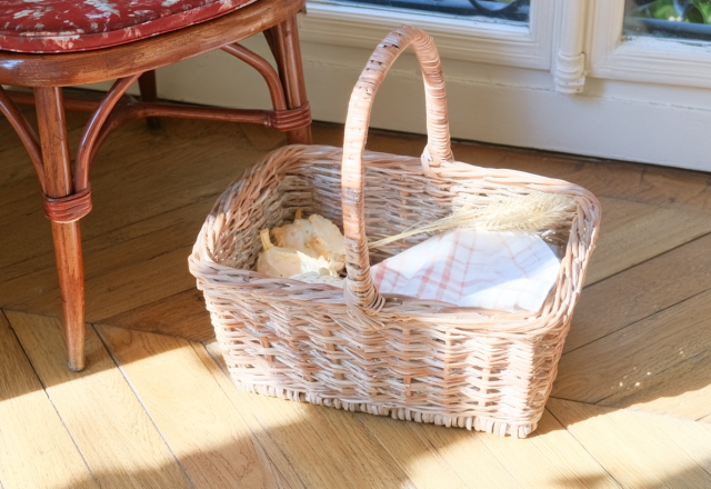 A wicker market basket with linen and dried wheat sitting on sunlit wooden floors next to an antique chair.