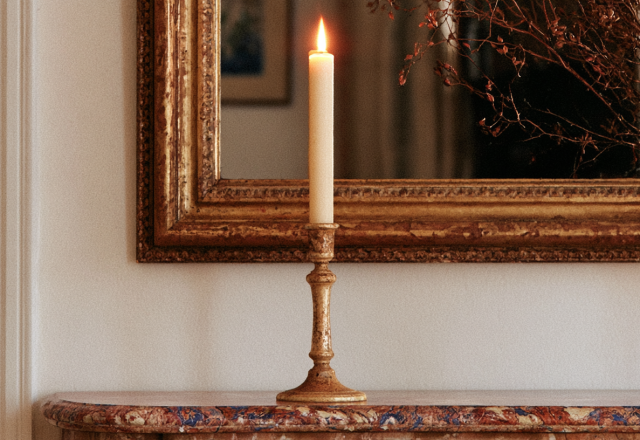 An antique brass candle holder with a lit taper candle resting on a marble console table.