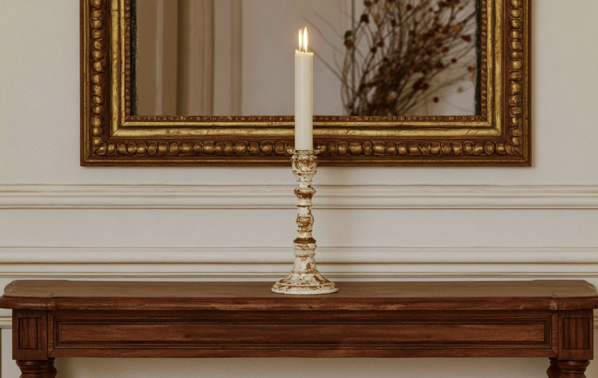 An antique brass candle holder with a lit taper sits on a wooden console table beneath an ornate mirror.