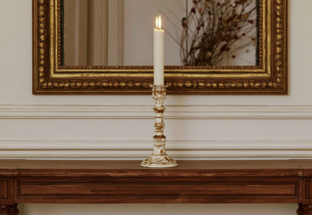 An antique brass candle holder with a lit taper sits on a wooden console table beneath an ornate mirror.