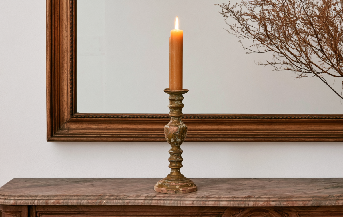 An antique brass candlestick holder with a burning candle on a marble mantel beneath a large wooden mirror.