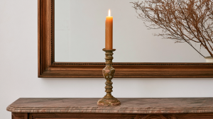 An antique brass candlestick holder with a burning candle on a marble mantel beneath a large wooden mirror.