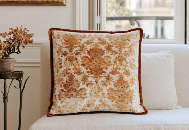 A patterned damask decorative throw pillow with terracotta trim resting on a white sofa next to a vintage plant stand.