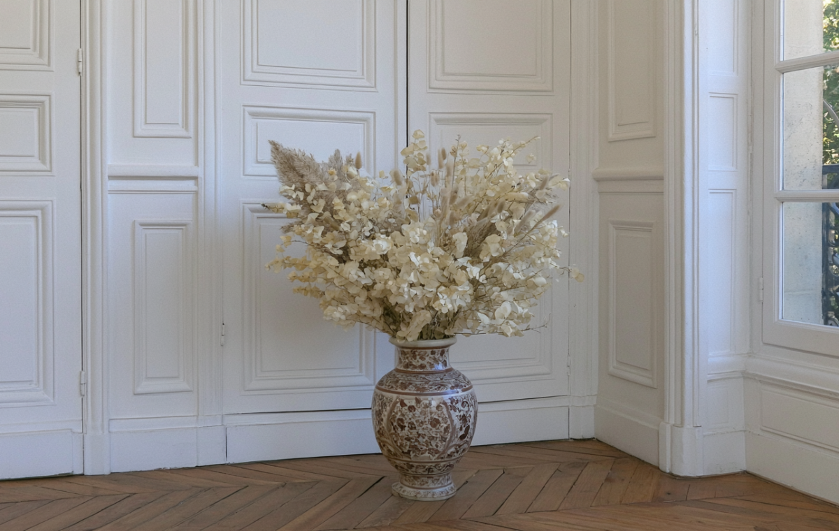 Dried floral arrangements in a patterned floor vase set against white paneled doors and parquet flooring.