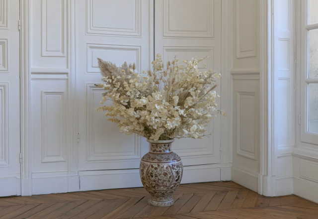 Dried floral arrangements in a patterned floor vase set against white paneled doors and parquet flooring.