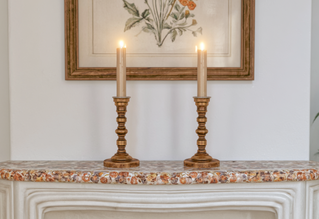 Two brass mantel candle holders with lit candles placed on a decorative marble fireplace shelf.