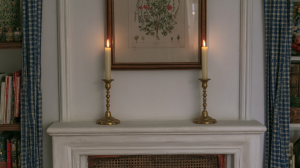 Two lit brass candlestick holders placed symmetrically on a white fireplace mantel beneath a framed botanical illustration.