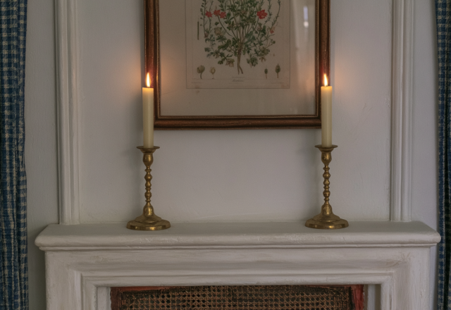 Two lit brass candlestick holders placed symmetrically on a white fireplace mantel beneath a framed botanical illustration.