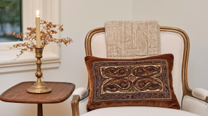 A brown velvet accent pillow with paisley pattern resting on an antique white chair next to a wooden side table with a candle.