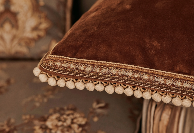 Close up of rich brown velvet throw pillow with gold brocade trim and pom pom fringe in a living room.