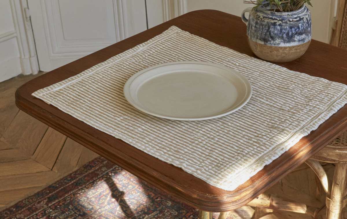 A minimalist striped placemat and ceramic plate on a wooden table in a sunlit breakfast nook.
