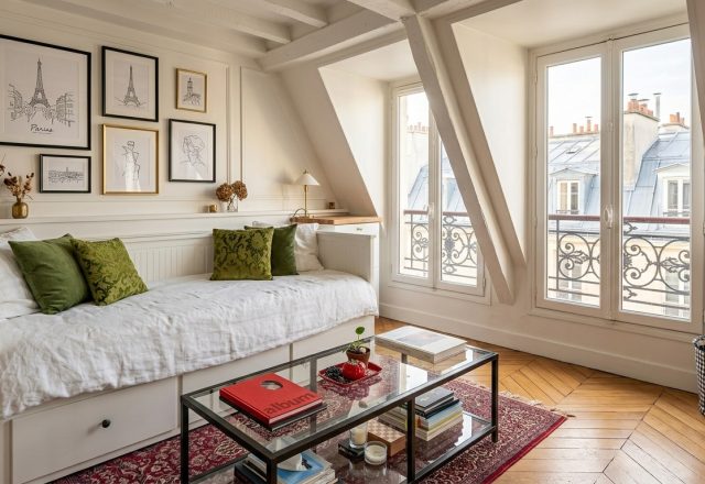 Bright attic living space featuring a white daybed, olive green cushions, and sloped windows overlooking classic rooftops.