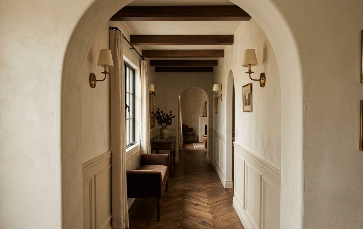 A warm transitional hallway featuring plaster walls, dark wooden beams, herringbone floors, and graceful archways.