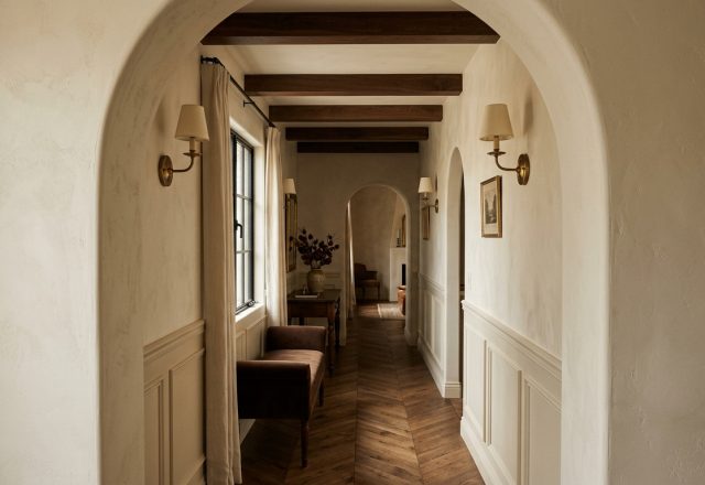A warm transitional hallway featuring plaster walls, dark wooden beams, herringbone floors, and graceful archways.