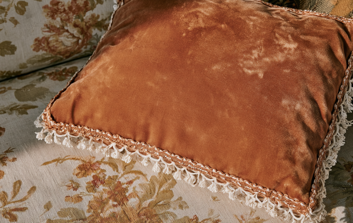 A rust velvet throw pillow with decorative fringe resting on a floral upholstered sofa.